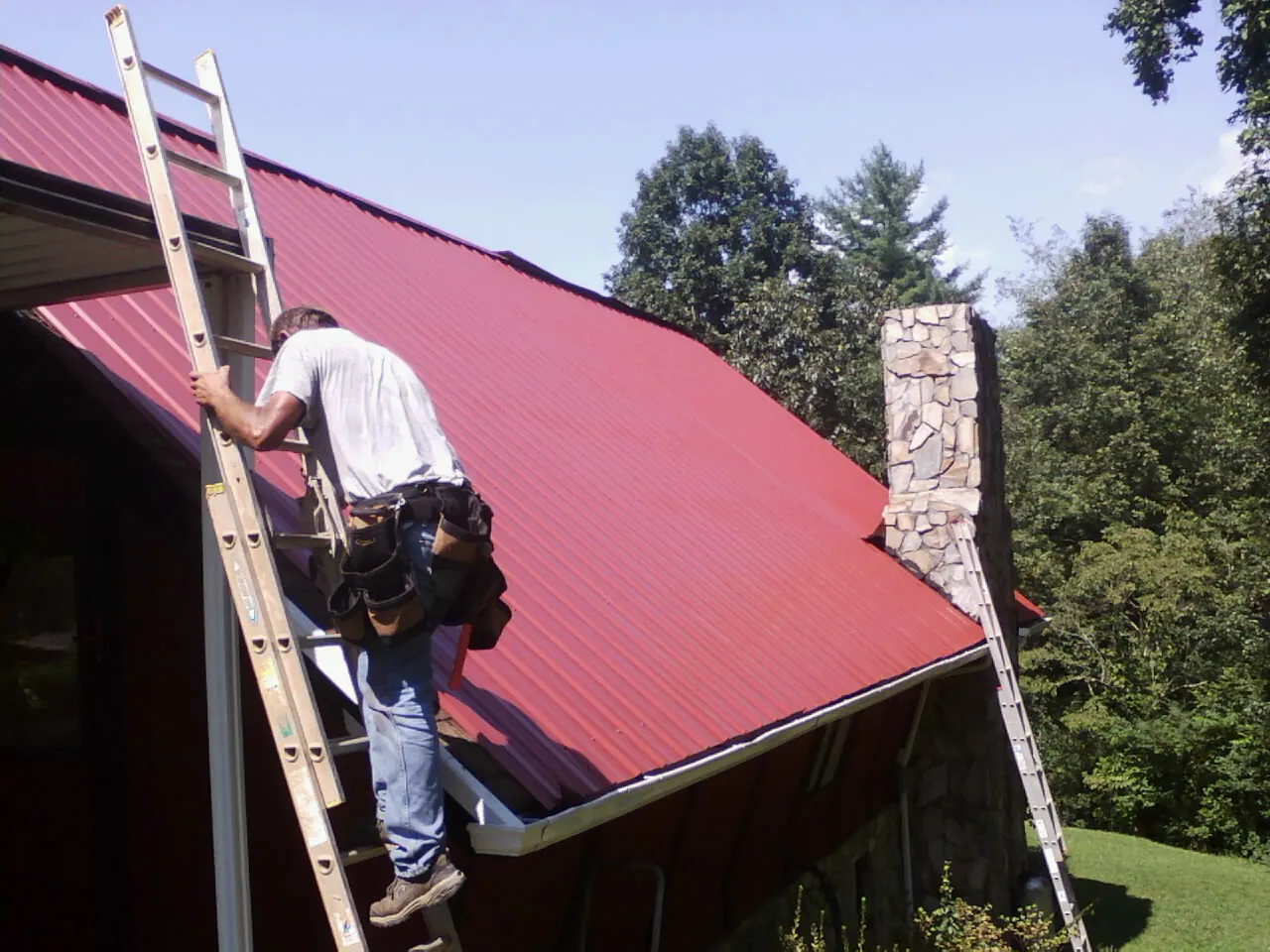 Roofer on a ladder beside a red metal roof and stone chimney — Soffit & Fascia Repair by Roofing Brothers in Morganza